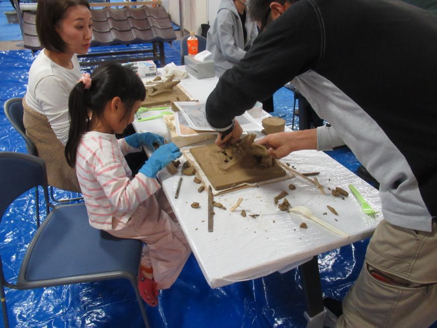 白いビニールが貼られた長机の椅子に女性と女の子が座り、粘土のようなもので作品をつくっている様子を写した写真