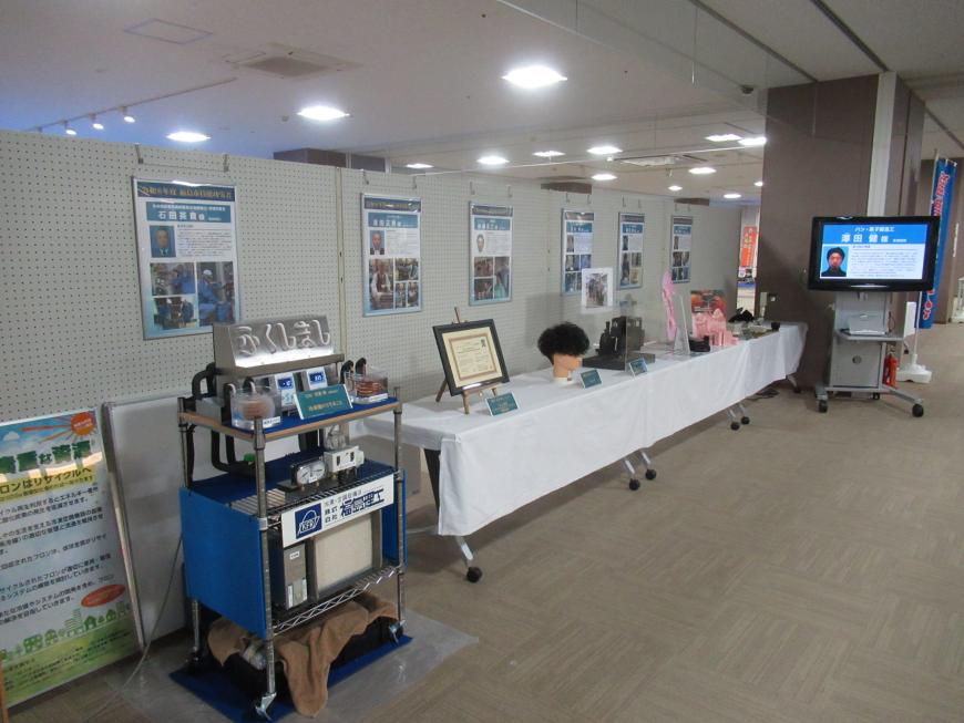 長机にいろいろな物が展示され、奥のモニターに人が映し出された技能功労者特設ブースを写した写真