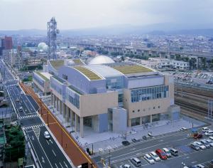 線路沿いの敷地内に駐車場が完備された、ベージュ色の外壁の福島市子どもの夢を育む施設こむこむの建物外観の写真