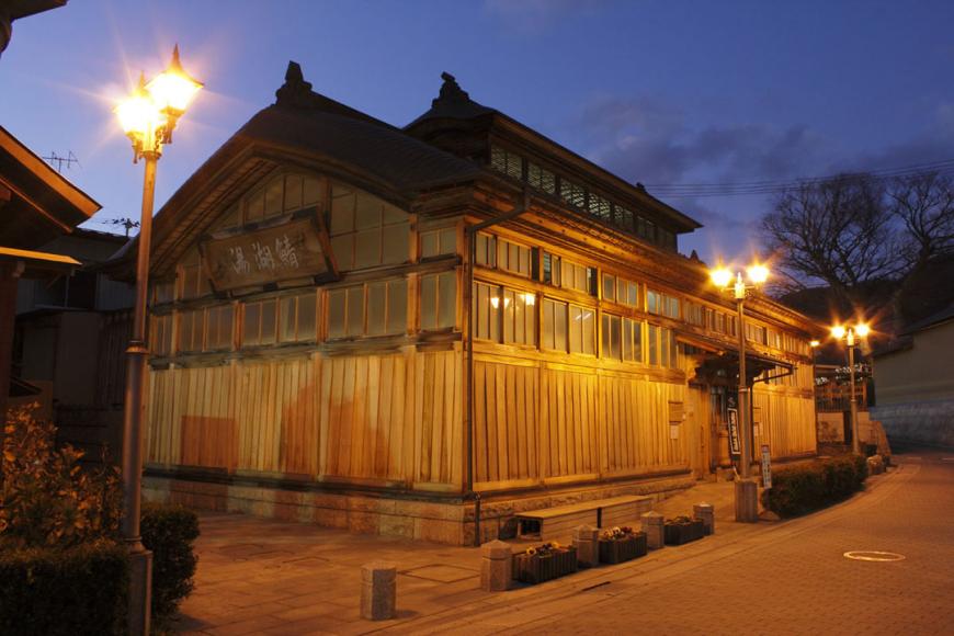 薄暗くなってきて街灯の光が灯った奥に建つ、日本最古の木造建築様式の鯖湖湯の建物外観の写真