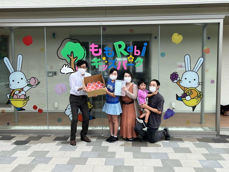「ももRabiキッズパーク」と書かれたガラス窓の前で、小さな男の子を抱っこした父親と母親、桃が入った段ボールを市長と一緒に持った女の子が並んでいる写真