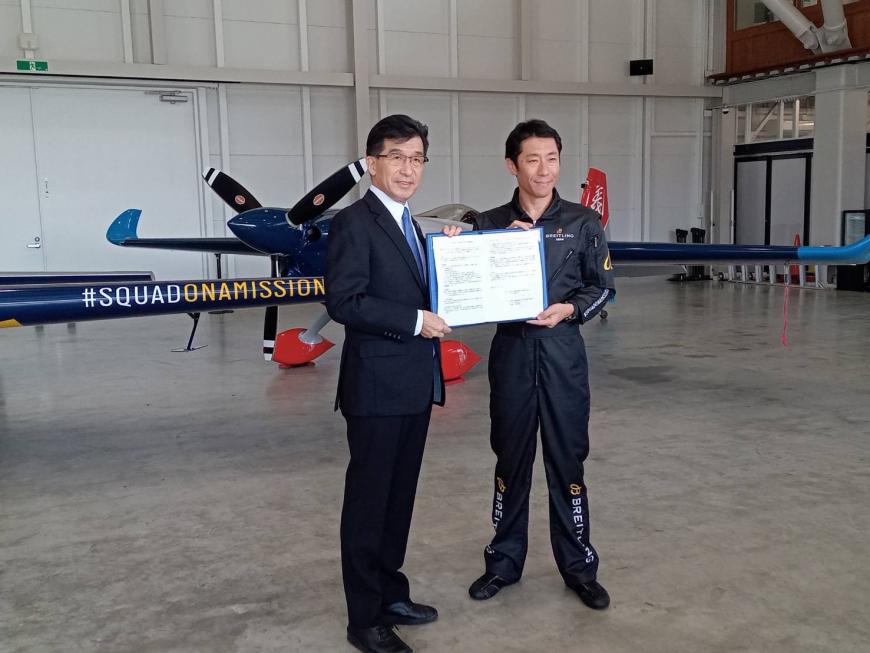小型飛行機が置かれた倉庫の中で、室屋義秀氏と市長が一緒に協定書を持ち並んでいる写真