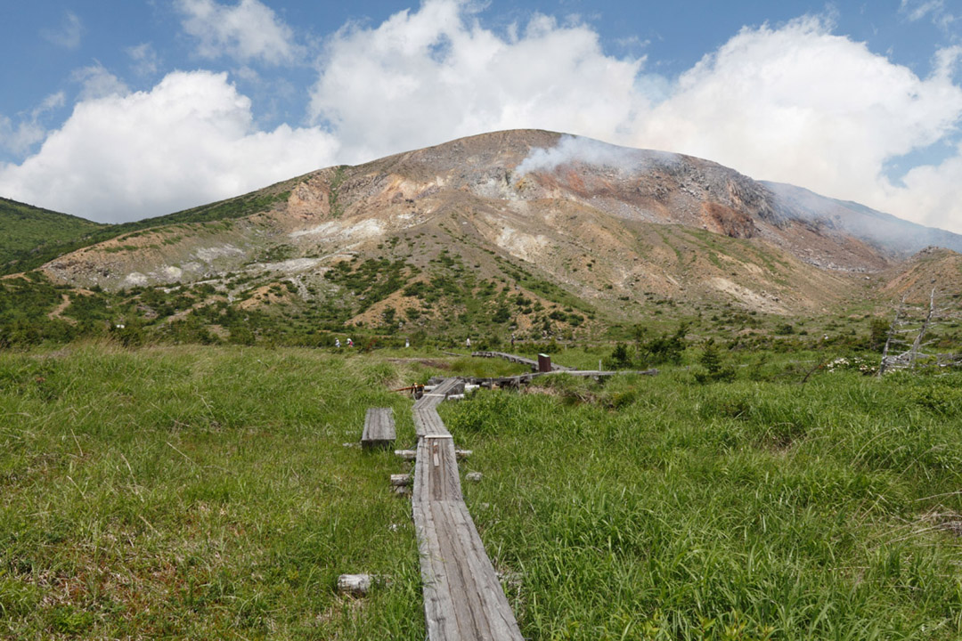 草むらの中に整備された木道の先に、もくもくとした煙が出ている一切経山の写真