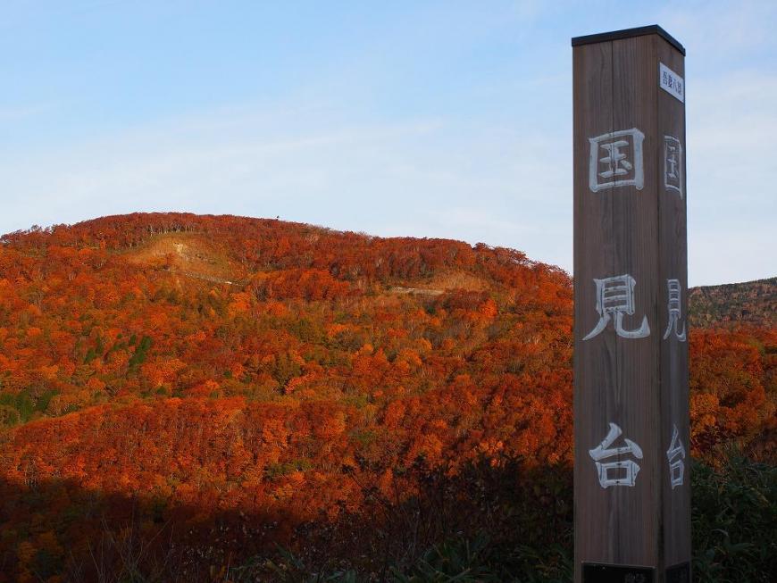 木が朱色に染まった山の手前に立つ国見台と書かれた標柱の写真