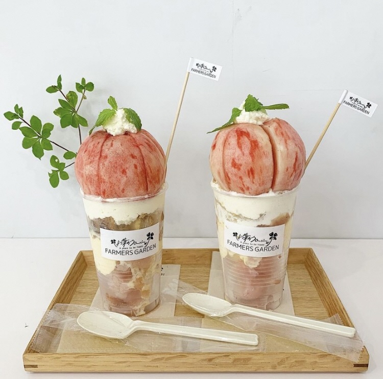 木製トレイの上に店の名前が書かれたラベルが貼られた透明の容器の上に桃がまるごと一個のっている2つのパフェの写真