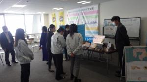商品が置かれた長机の前に数名の女性が並び、女性担当者から説明を受けている株式会社環境分析研究所のふくしまチャレンジ・フィールドの様子の写真