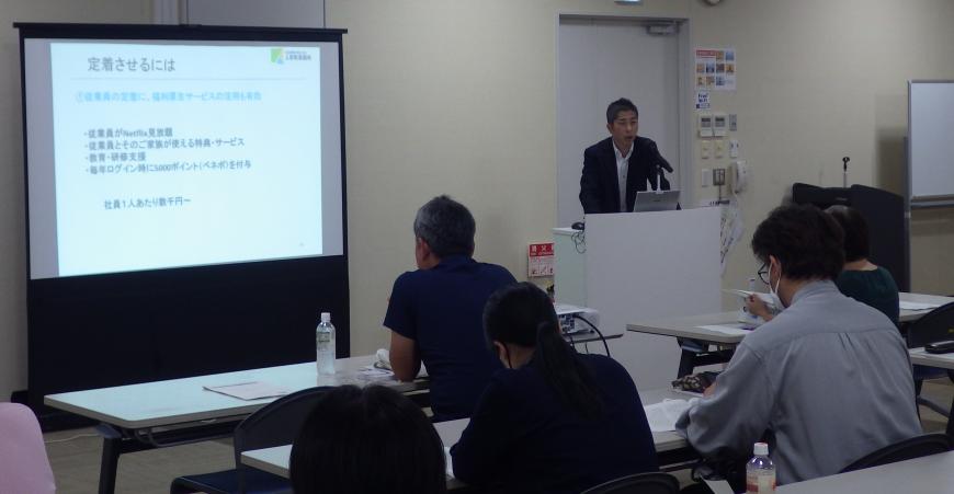 前方に設置されたスクリーンに資料が映し出され、右側の檀上に立つ男性の話を参加者が熱心に聞いている写真