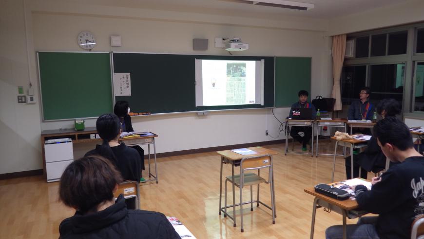 資料が映し出された黒板の右側に2名の男性担当者が座り、自席に座った生徒たちが熱心に話を聞いている様子を後方から撮影した写真