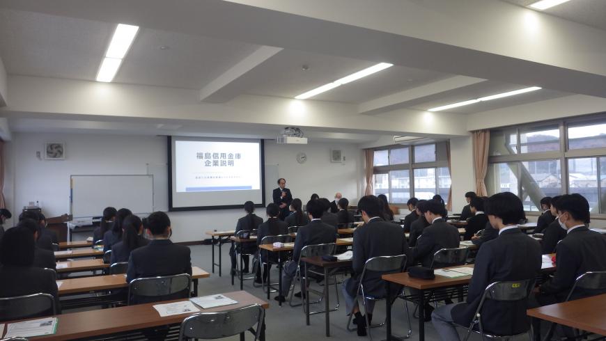 スクリーンの右側に立つ男性担当者の話を、長机の席に2名ずつ座った高校生が熱心に聞いている様子を左後方から撮影した写真