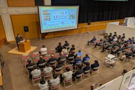 前方中央に資料が映し出された大きなスクリーンが設置され、左側に設置された壇上に立つ市長の話を会場に設置された椅子に座った参加者の方々が聞いている写真 1booth パネル展示されたブースの担当者に話を聞いている参加者の写真