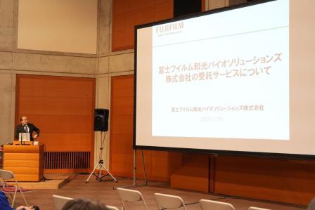 「冨士フィルム和光バイオソリューションズ株式会社の受託サービスについて」と書かれた資料が映し出されたプロジェクタースクリーンの左側に設置された演台に立ち、男性が話をしている写真
