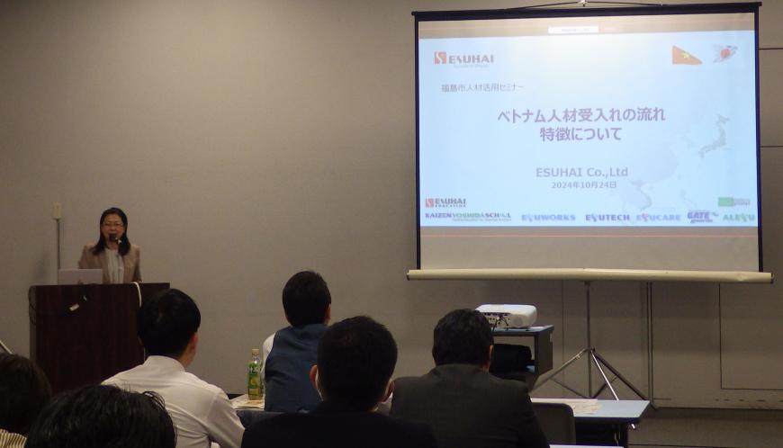 前方に設置されたスクリーンに資料が映し出され、左側の檀上に立つ女性の話を参加者が聞いている様子を後方から撮影した写真