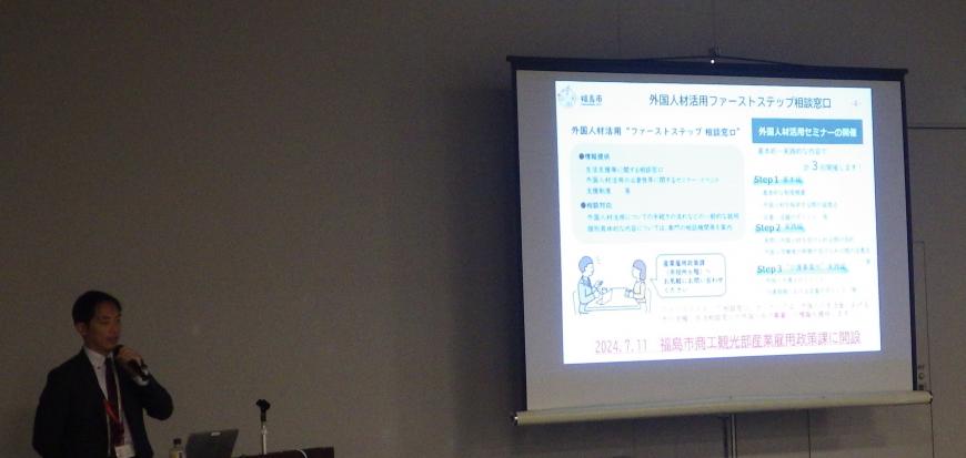 前方に設置されたスクリーンに資料が映し出され、左側の檀上に立つ男性が話をしている写真