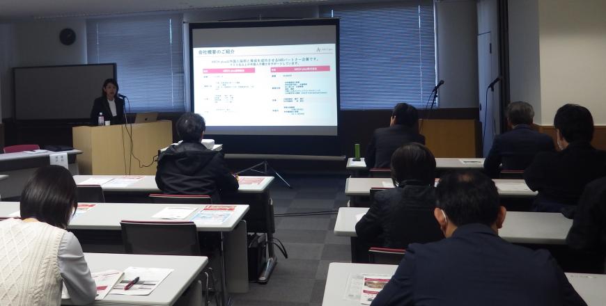 前方に設置されたスクリーンに資料が映し出され、左側の檀上に立つ女性の話を聞いている参加者を後方から撮影した写真