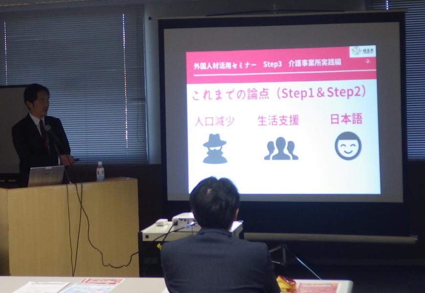 前方に設置されたスクリーンに資料が映し出され、左側の檀上に立つ男性が話をしている写真