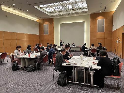 前方に立つ担当者が資料を手に持ち、各テーブルの参加者の人達もその資料に目を通して話を聞いている交流会の写真