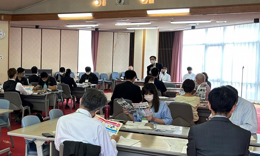 机に座っている人達がチラシを手に持ち前方に立っている男性の話を聞いている交流会の写真