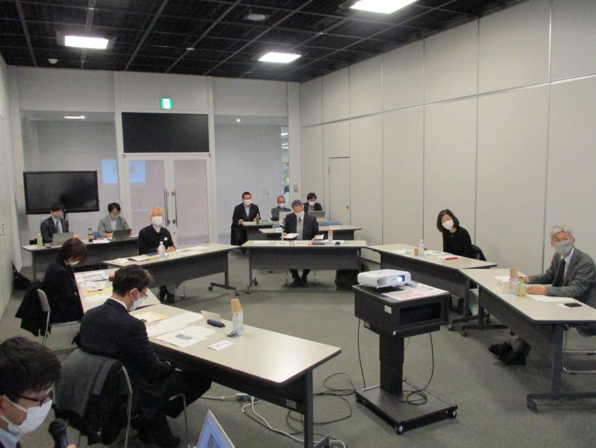 中央のプロジェクターを囲むように設置された長机の席に参加者が座り、プロジェクターで映し出されたスクリーンを見ている会議の様子の写真