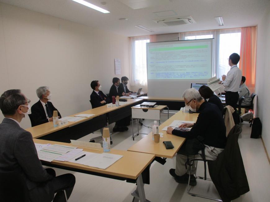 前方に資料が映し出されたスクリーンが設置され、右端に立っている男性の話をコの字型につなげられた長机の席に座った参加者が熱心に聞いている写真