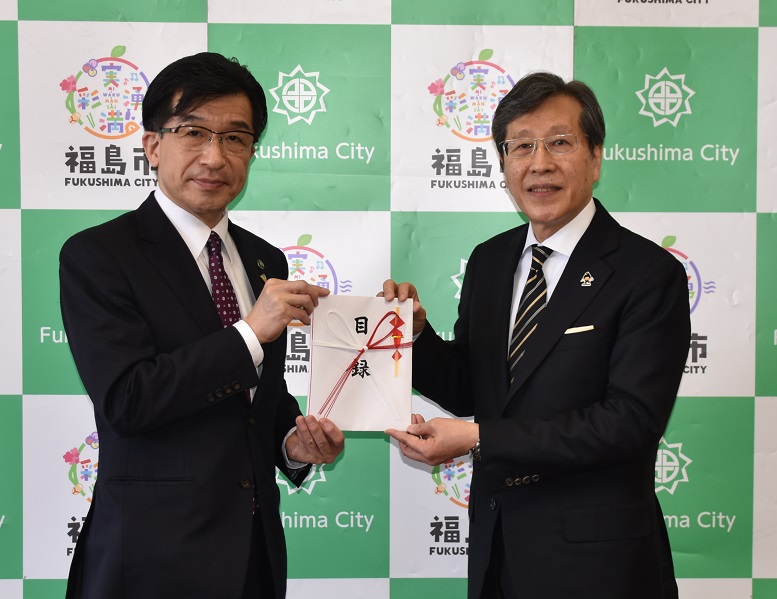 福島市のバックパネルの前で、浅石理事長と市長が一緒に目録を持ち並んでいる記念写真