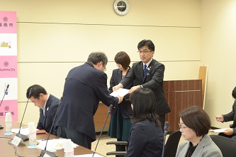 自席から立ち上がった男性が、市長から委託状を受け取っている交付式の様子の写真