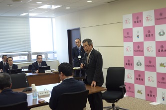 机の端の席の男性が立ち上がってマイクを手に持ち話をしているのを参加者が聞いている会議の様子の写真