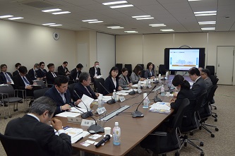 前方のスクリーンに資料が映し出され、参加者が手元に資料を見ながら話しを聞いている会議の様子の写真