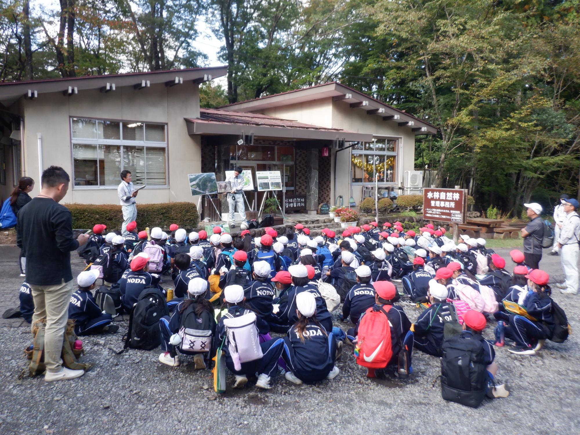 3枚のパネルの前に立って森林の役割の説明をしている男性としゃがんで話を聞いている子供たちの写真