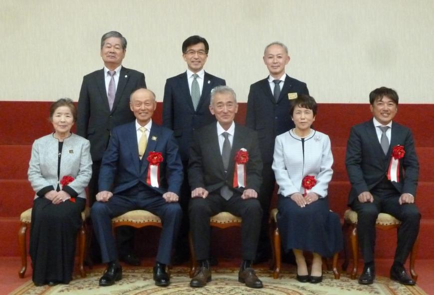 朱色の絨毯が敷かれたひな壇の前の前列の椅子に赤い胸章リボンを付けた受賞者の5名と、後列に市長と2名の関係者の男性がが立っている記念写真