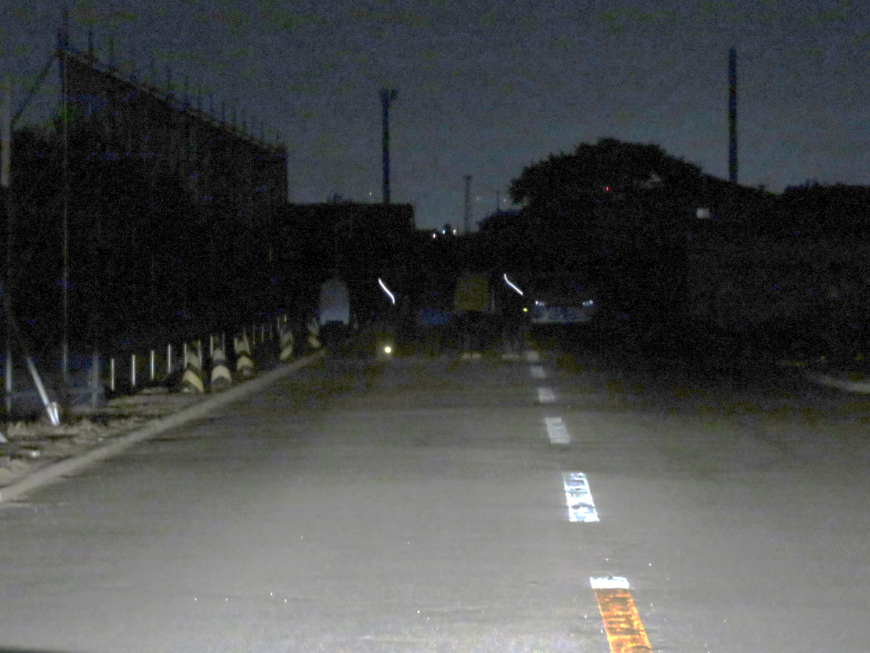 夜間、ライトの明かりの先に歩行者と車がうっすらと見える中、歩行者が着用したたすきの反射材が白く反射している様子の写真