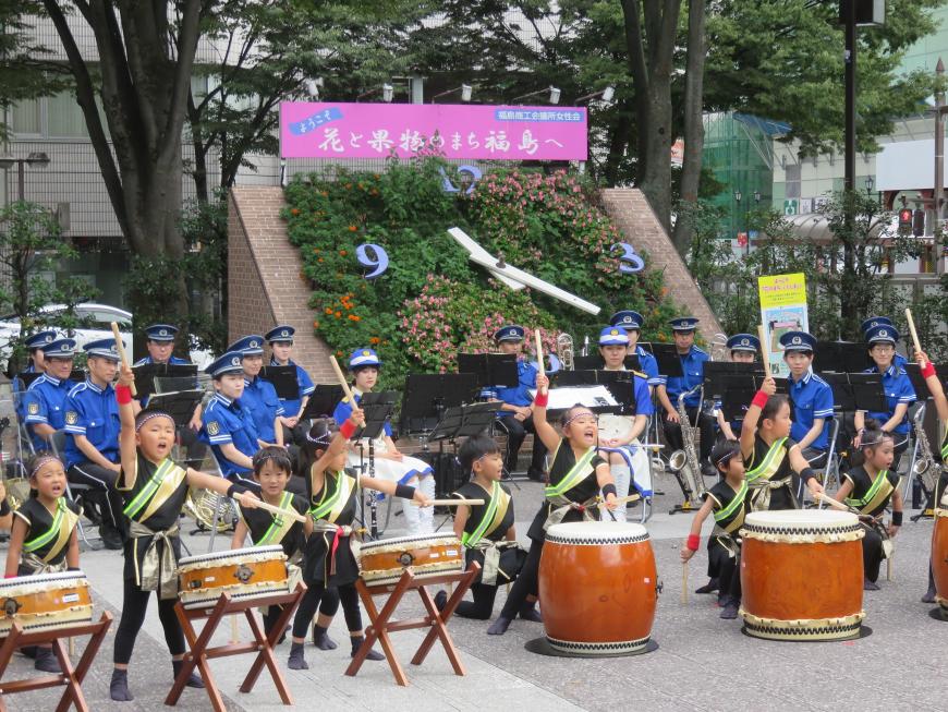 駅前広場の花時計前で、警察の吹奏楽団が座る前に置かれた太鼓を園児たちが演奏している様子の写真