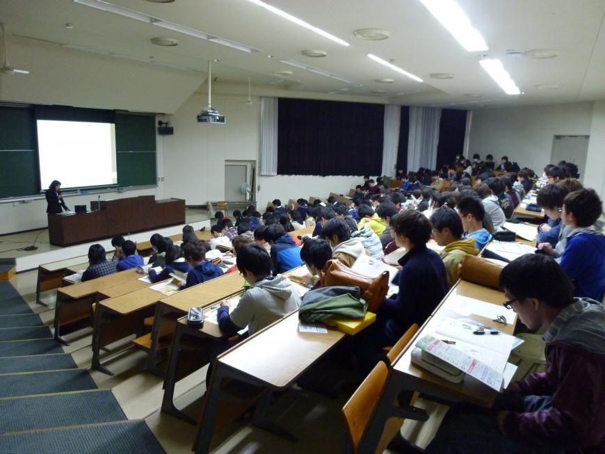 階段状になった席に大勢の学生が座り、スクリーンが設置された黒板前に講演者が立っている出前講座の様子の写真