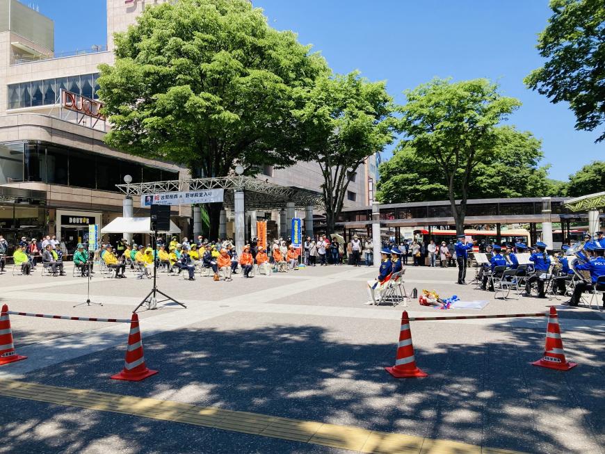 駅前広場の右側で警察音楽隊が演奏をし、左側に並んだ椅子にオレンジや黄色、緑のジャンバーを着た関係者たちが座っている様子の写真