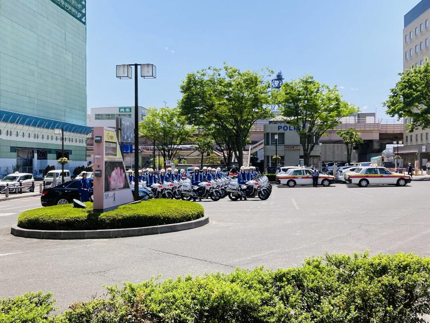 駅前ロータリーの駐車場にたくさんの白バイが並び、白バイ隊がバイクの横に整列している写真