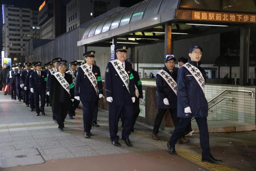 白いたすきをかけた内堀知事に続き、制服を着た大勢の警察官が2列になって歩道を歩いている様子の写真