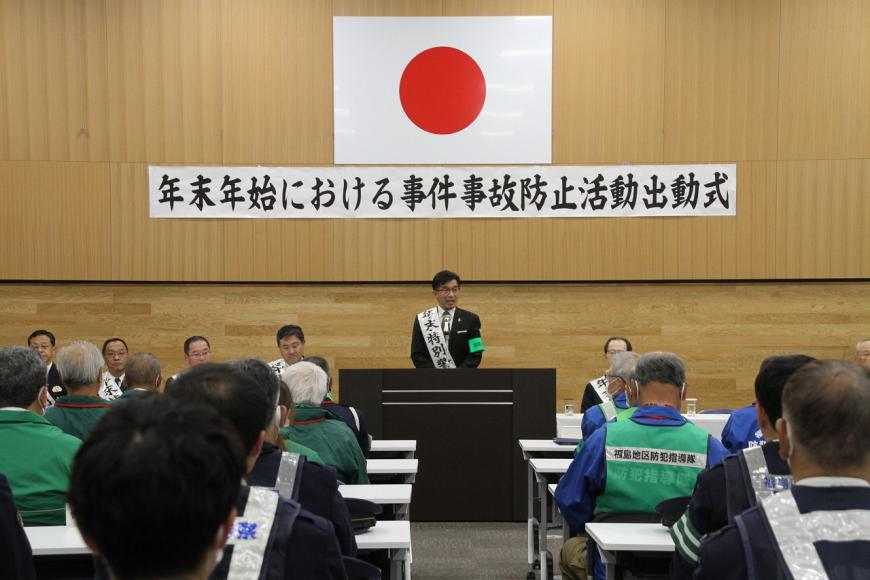 日本国旗と「年末年始における事件事故防止活動出動式」の横断幕が掲げられた壁の前の演台に立った市長が、長机の席に座った出席者たちに話をしている写真