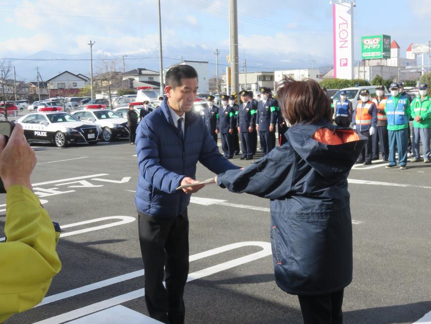駐車場に複数のパトカーが停まり、たくさんの警察官や関係者が整列している前で、女性が男性に「交通安全啓発はし袋」を贈呈している様子の写真