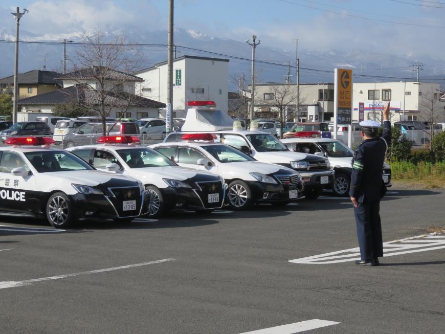 整列して停まったパトカーなど5台の警察車両の方を向いて前に立った警察の制服を着た男性が右手を挙げている写真