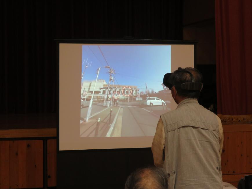 道路が写ったスクリーンの前にVRシミュレーターを装着して立つ男性の写真