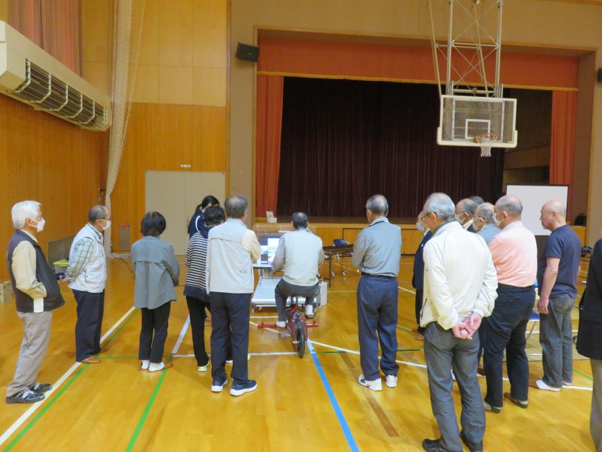 自転車VRシミュレーターを体験する高齢者男性を、周りに集まったたくさんの高齢者たちが見ている様子の写真