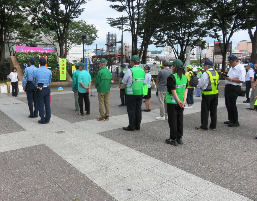 まちなかの広場に集まった警察官や蛍光色のビブスを着た職員たちが整列して立っている様子を後方から写した写真