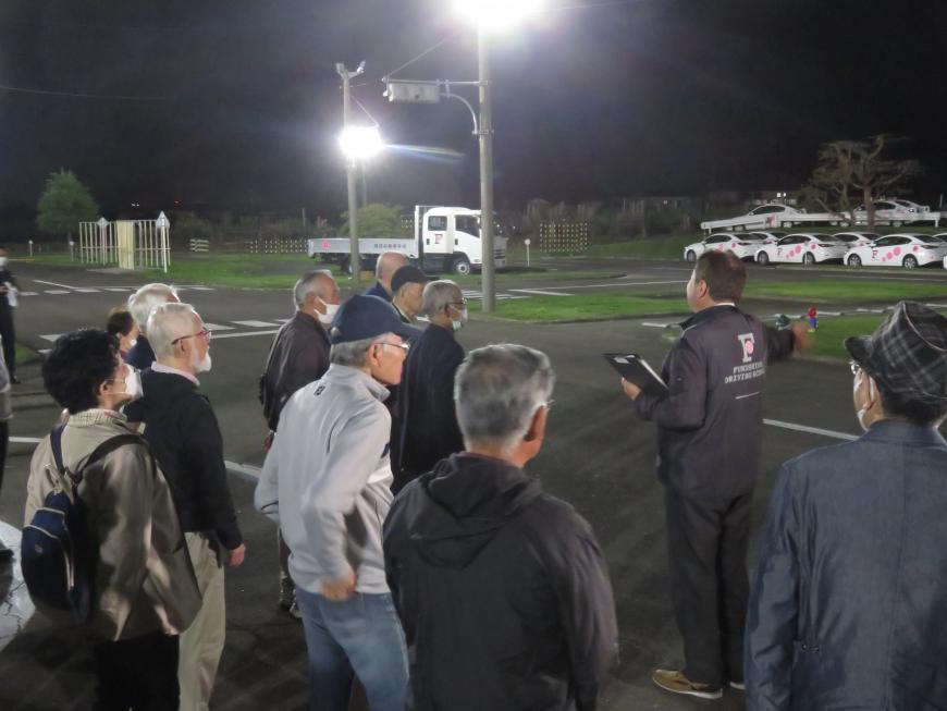 大勢の高齢運転者が夜間に自動車学校の敷地内で職員の話を聞いている様子の写真