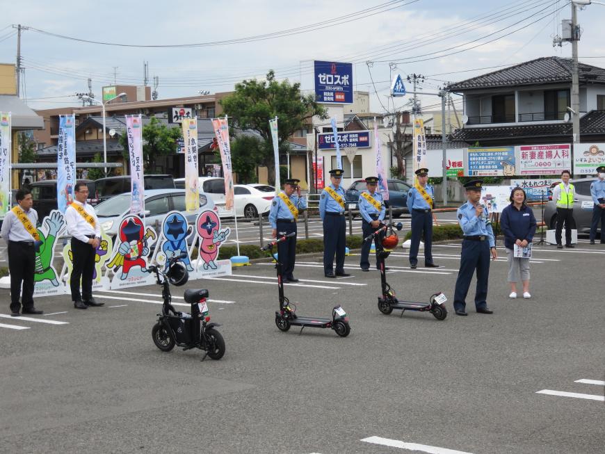 黄色のたすきをかけた2人の関係者と4人の警察官が並ぶ前に3台の電動キックボードが並び、その右側に立った警察官の男性がマイクで話をしている様子の写真