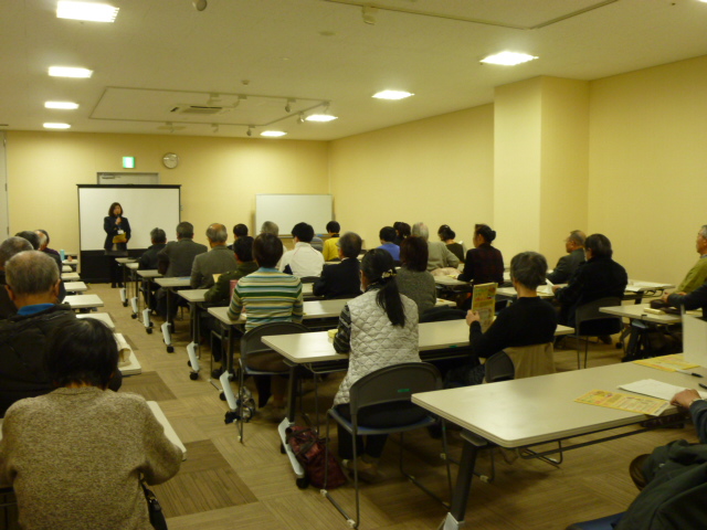 会議室に横3列でたくさん並んだ白い長机の席に、多くの参加者たちが着席し正面中央の大きなスクリーンの前に立ち話をしている女性の方を向いている写真
