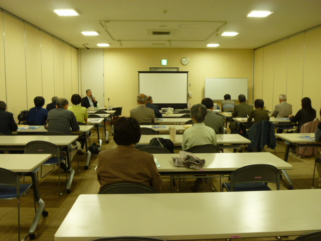 会議室に横3列でたくさん並んだ白い長机の席に、参加者たちが着席し正面中央の大きなスクリーンの方を向いている写真