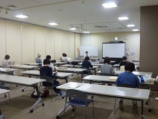 前方にホワイトボードとスクリーンが並べて設置された会議室に白い長机がたくさん並べられ、出席者たちが席についている研修会の写真