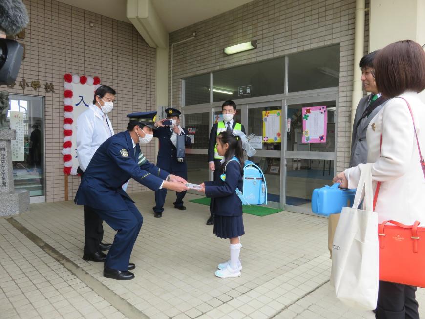 小学校の入り口前で、腰を屈めた警察官の男性が、紺色の制服を着て、水色のランドセルを背負った女の子にパンフレットを手渡している様子の写真