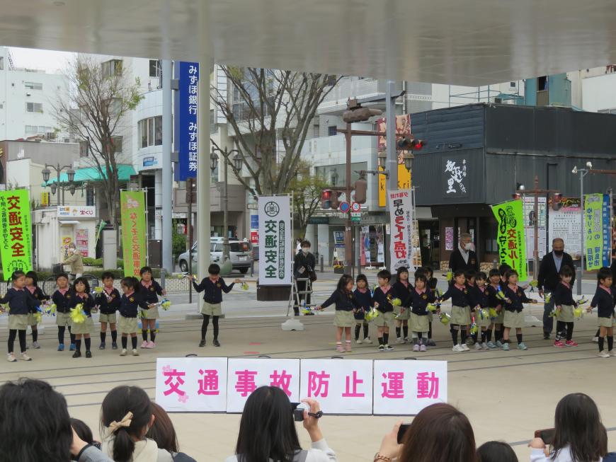 屋外の屋根付きスペースで、「交通事故防止運動」と書かれた看板の後ろに並んだおそろいのユニフォームを着たたくさんの園児たちがダンスを披露している様子の写真