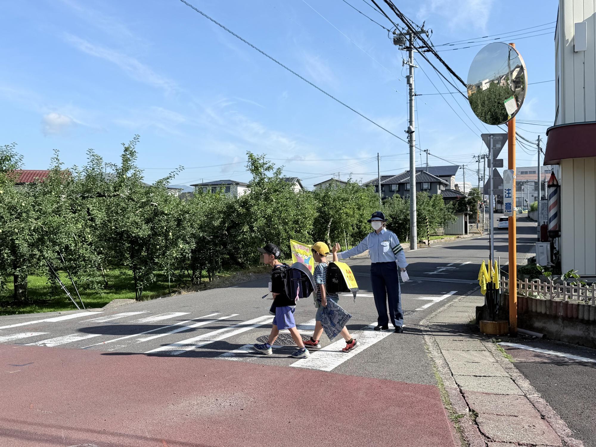 制服を着用した交通教育専門員が、横断旗を掲げて横断歩道上に立ち、横断歩道を渡る児童の安全を確保している様子