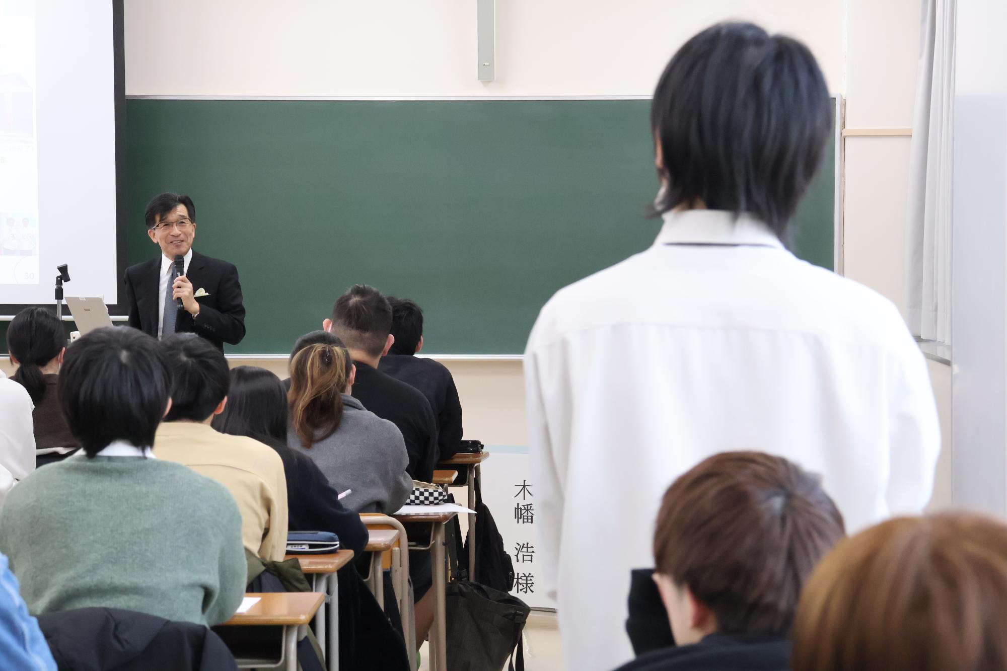 黒板の前に立っている市長と、自席から立ち上がった白いシャツを着た人が話をしている写真